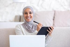 Close-up photo. Muslim young woman in hijab freelancer work from home online. Sitting on the sofa with a laptop and a folder with documents. He writes, fills in with a pen, smiles at the camera. photo