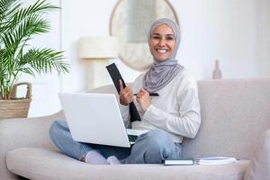 Young female student of oriental origin in hijab studying from home. Sitting on the sofa in the lotus position with a notebook and writing with a pen in a notebook, folder. Smiling at the camera. photo