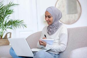 Distance Learning. Young Latin American woman in hijab student studying online. He sits at home on the sofa with a laptop on his lap and writes down a lecture, lesson, course in a notebook. photo