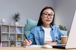 Job interview. A young beautiful Asian business woman in glasses sits at a desk, conducts an interview in the office, hires, writes down a questionnaire in a notebook, interviews the interlocutor. Hello, looking at the camera. photo