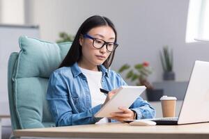 Young Asian woman studying online while sitting in office, writing lecture notes in notebook, using laptop for studying. photo