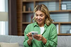 Joyful senior mature woman at home smiling and sitting on sofa, using phone in living room, browsing online pages and dialing message call. photo
