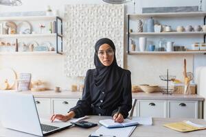 Successful and confident young woman in black hijab and burqa sitting at home at kitchen table and working remotely with documents and laptop. He looks seriously into the camera. photo