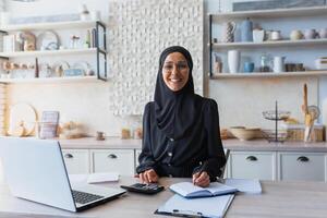 Portrait of young Muslim woman in hijab and burqa working as accountant, auditor from home. Remotely works on a laptop, with documents and a calculator. Smiling at the camera. photo