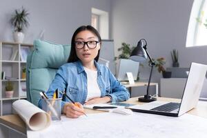Portrait of a young beautiful Asian woman architect in glasses working at a desk at a computer and drawing on a project. Looks into the camera photo