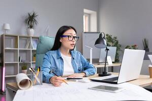 Young beautiful Asian woman architect designer wearing glasses working at desktop computer and drawing with pencil, working on project. photo