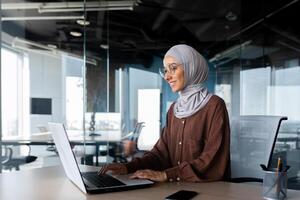 Young beautiful muslim woman in hijab working inside modern office, businesswoman using laptop at work, typing on keyboard while sitting at workplace smiling and happy with achievement result. photo