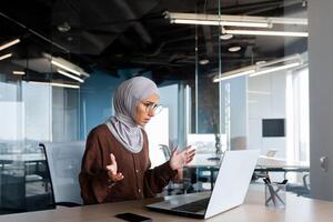 A young Arab woman in a hijab is sitting at a desk in the office and working on a laptop. Problems with the Internet, the computer is broken, he looks worriedly at the monitor, spreads his hands photo