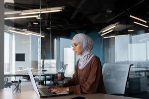 Serious and thinking woman in hijab working inside office with laptop, Muslim female programmer typing on keyboard writing software code at workplace working artificial intelligence development. photo