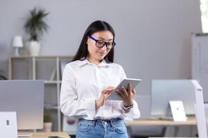Young beautiful Asian teacher working with tablet, in classroom with computers at school, woman in white shirt and glasses reads from tablet photo