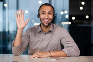 Smiling joyful African American man in headset talking on call, consulting clients in modern office, working in online store support, waving at camera, camera view. photo