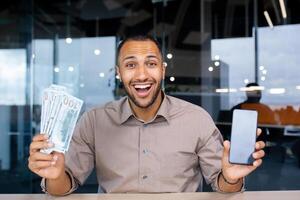 Happy bearded man in office holding stack of dollars and showing phone screen to camera, happy with winning, online earnings, looking at camera smiling. photo