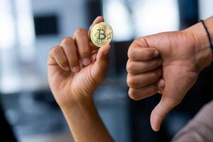 Close up portrait of hands holding bitcoin virtual currency and showing dislike thumb down, office space. photo