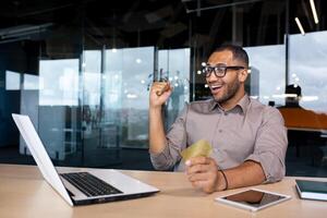 Happy African American man in glasses sits in the office in a co-working space at a table in front of a laptop, holds a card in his hands, makes online purchases, pays on the Internet. photo