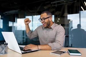 Successful Indian programmer celebrating successful completion of testing new software, man happy with achievement results working with laptop, inside office, online winner. photo