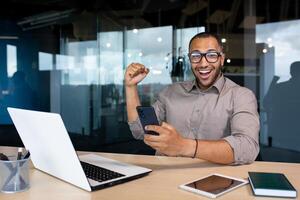 Joyful Arab businessman holding a phone in his hand and joyfully raising his hand up, sitting at a table in a foyer, happy with good news, smiling and looking at the camera. photo