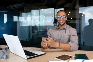Portrait of a young successful businessman, the man is smiling and looking at the camera, holding a phone in his hands, using an application on a smartphone, working inside the office with a laptop. photo