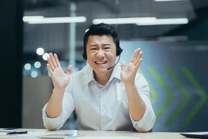 Portrait of an angry young Asian man sitting in the office at a ,desk wearing a headset and expressively begging on a call, while looking at the camera. photo