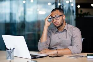Frustrated and sad man at workplace reading online message from laptop, businessman sitting inside office at workplace, desperate unhappy, got bad news, bankruptcy, layoff. photo