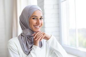 The photo is a close-up portrait of a young beautiful muslim woman in a hijab standing inside by the window, leaning her face on her hands and smilingly posing for the camera.