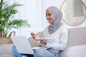 Arab young female student studying online at home via laptop. Sitting on the sofa in the lotus position, writing in a notebook, communicating via call. photo