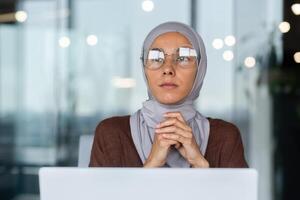 Close-up photo. Portrait of a young beautiful Arab student woman in a hijab. Sitting at the table, working, studying at the computer. He looks thoughtfully and seriously to the side, to the window. photo
