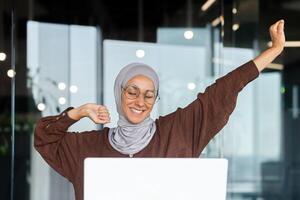 Portrait of young Arab businesswoman in hijab and glasses sitting in office at table with laptop. She is tired, has finished her work day, has done her work, stretches her hands up, smiles, rests. photo