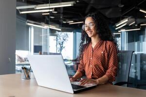 Cheerful and successful indian woman programmer at work inside modern office, tech support worker with laptop typing on keyboard smiling. photo