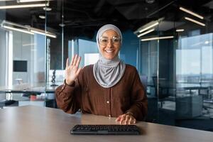 Young Muslim female student sits in a co-working space in front of the camera and greets with a hand, goes through an interview and presentation, talks on a call. photo