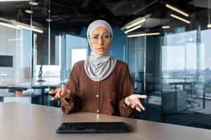 Serious young Arab woman sitting in the office in front of the camera at the table and talking, explaining, having a discussion, solving questions on an online conference. photo