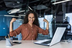 Portrait of a strong business woman female worker working inside office building, sitting a desk and using a laptop, smiling and looking at the camera holding hands up gesture superpower and success photo