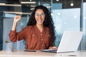 Portrait of a successful hispanic businesswoman working inside a modern office building smiling and looking at the camera, holding her hand up in a gesture of success and triumph. photo
