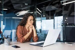 Pensive and focused female programmer web developer working in modern product development company Hispanic woman sitting and thinking using laptop to write code. photo