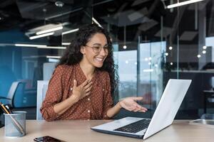 Happy hispanic businesswoman working in modern office using laptop for call and online meeting with fellow employees, woman smiling and having fun giving a presentation photo