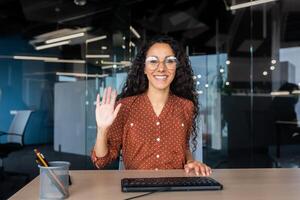 Portrait of a young Latin American female student. Sits in front of the camera, waves, greets, talks via call, smiles. Sitting at the desk in the office. photo