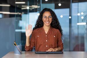 Webcam view, happy and smiling Latin American woman smiling and looking at camera waving greeting gesture, tech support and customer service worker, from online help. photo
