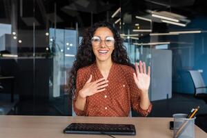 Online customer support call, Latin American woman looking at web camera smiling and consulting customers, helpline worker working inside office. photo