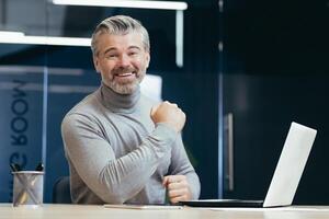 Portrait of a senior gray-haired successful businessman. A happy man is sitting in the office at the table with a laptop. Celebrates victory, success, smiles at the camera, shows well done. photo