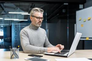 Senior and experienced programmer works in modern office at laptop, male web designer developer in glasses writes code programs new application, mature businessman works at desk. photo