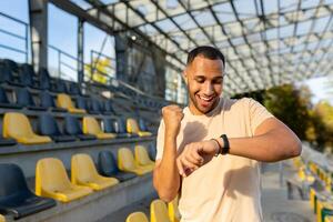 Happy and successful man satisfied with the results of physical training celebrating good results of fitness and running looking at smart watch, fitness bracelet walking in the air in the stadium. photo