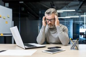 Sick mature businessman working in modern office, senior experienced man having severe headache, manager massaging temples while sitting on chair, manager using laptop for work photo