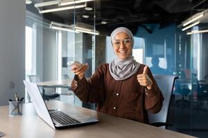 Portrait of a young Muslim woman in hijab working in the office, sitting at the desk and turning on the air conditioner with the remote control, pointing super finger and smiling at the camera. photo