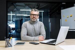 Portrait of senior programmer, mature team leader serious and focused looking at camera, businessman boss working inside office building, using laptop. photo