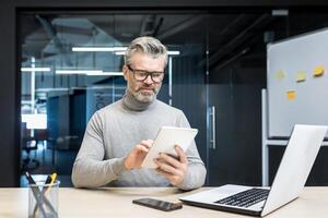 Senior experienced gray haired man in glasses using tablet computer, businessman at work inside modern office building photo