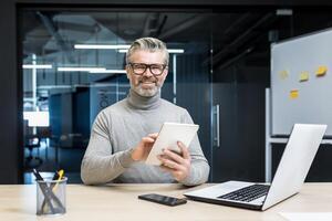 Portrait of successful programmer team leader, mature gray-haired engineer testing new app on tablet computer, businessman working inside modern office building, smiling and looking at camera photo