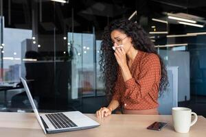 Sick woman working in office, hispanic woman having flu and cold drinking hot tea sneezing and coughing at workplace, using napkins, sitting at table with laptop. photo