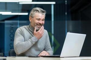 Mature and experienced senior gray-haired businessman working in modern office, watching fun and funny on laptop, company owner smiling and happy in work break. photo
