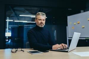 Portrait of serious gray haired boss, man looking at camera thinking, mature businessman inside office working at desk using laptop at work. photo