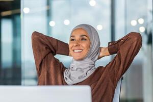 A dreamy and smiling young Arab woman in a hijab is sitting in the office on a chair, hands behind her head, resting. A successfully completed case, the end of the working day, plans for the future. photo
