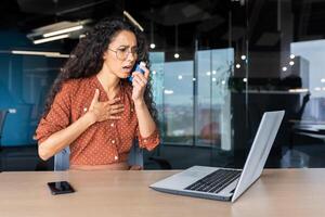 Sick woman at work having breathing problems, Hispanic female worker using inhaler with medicine for breathing relief and asthma, working inside office with laptop. photo